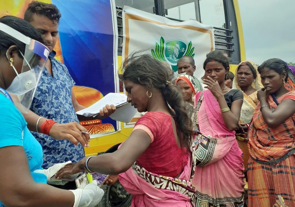 Root staff member Lucy wearing a plastic shield face covering and taking a villager's finger print while village representative ravi oversees it clipboard in hand and a small group of villagers look on.
