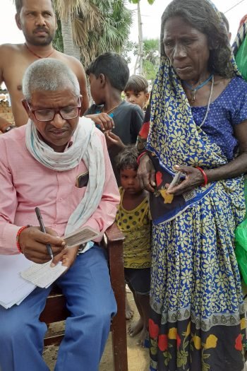 Vijay is seated on a chair with paper on his lap and a pen in his hand and examining a card held in his hand as a villager stands next to him watching.